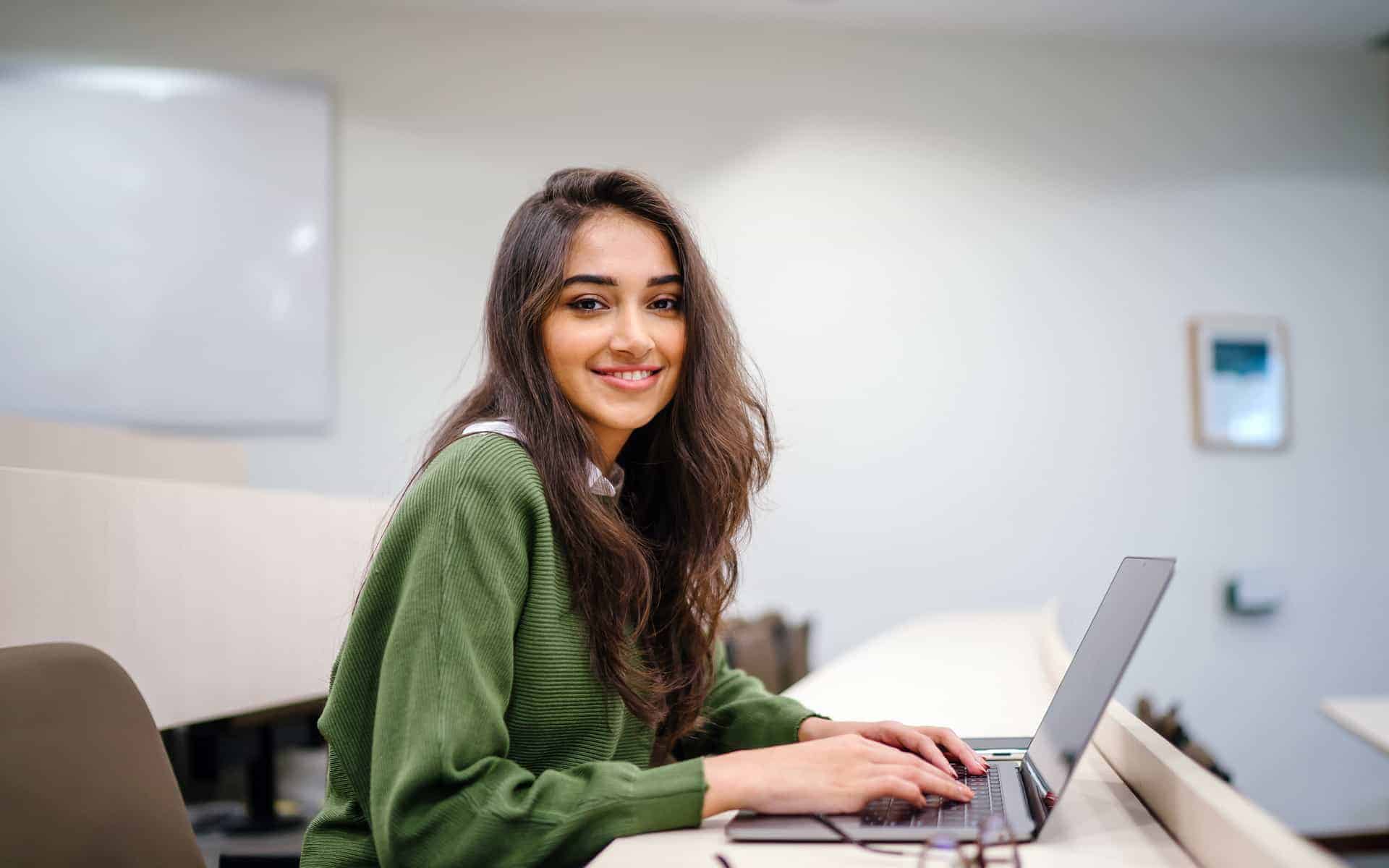 Retrato de una bella, joven e inteligente estudiante india asiática con un pantalones blanco y un rastreador verde sonriendo mientras trabaja en su laptop en un salón de clases de la universidad.
